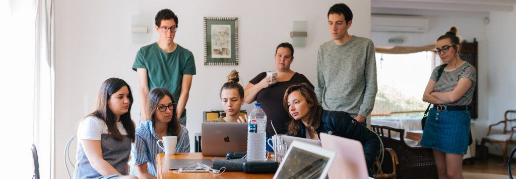 Four students standing behind four seated students at a table. One of the seated students is showing the rest of the group work on a laptop opened on the table.